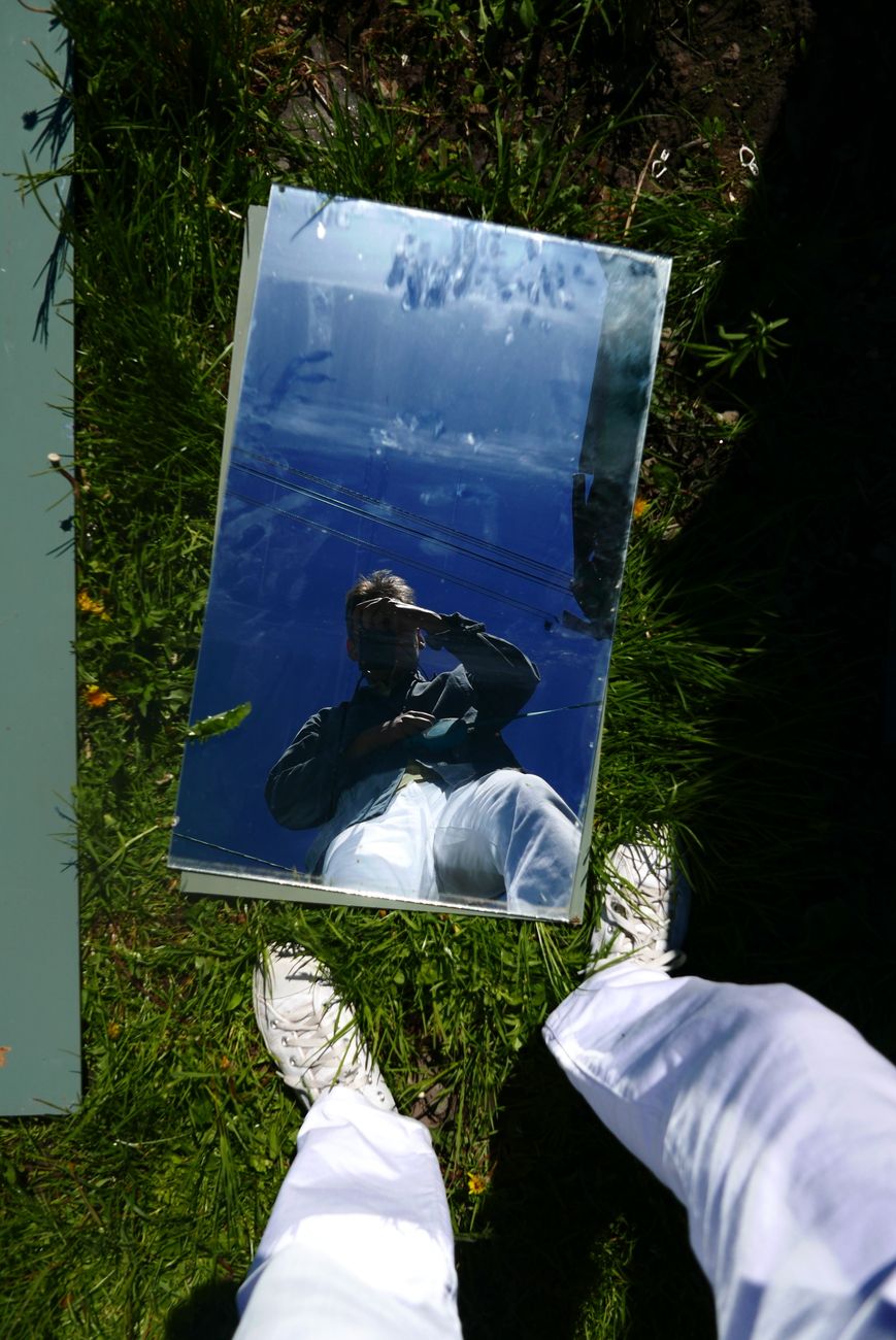 Reflet d'une personne dans un miroir posé sur l'herbe, tenant un smartphone et portant des vêtements blancs.